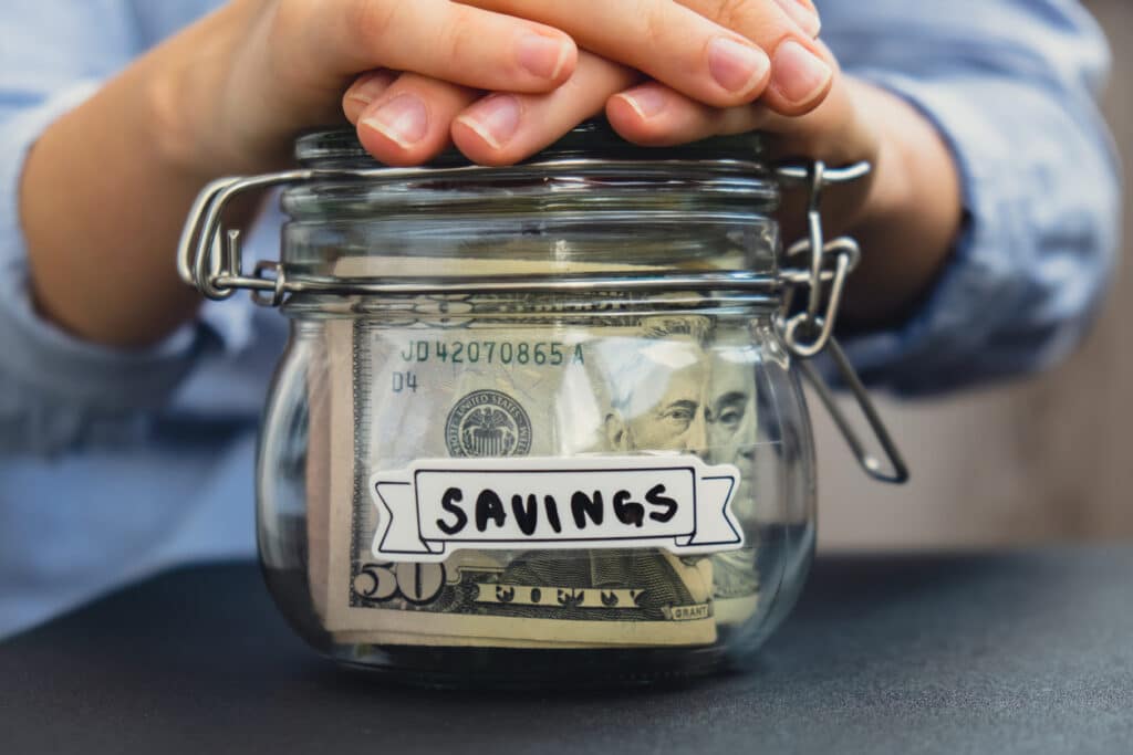 Hands holding glass jar with cash in it, labeled "Savings"